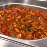 Greek-Style Chicken Stifado with Red Wine Sauce in a stainless steel foodservice hot bar insert. Braised chicken thigh pieces with red and green peppers and onions in a rich red wine sauce, ready for heat-and-serve foodservice programs